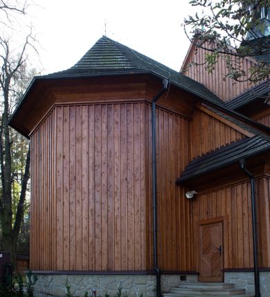 Zdjęcie nr 38: Kościół drewniany, orientowany, jednonawowy, wzniesiony na niewielkiej ceglanej podmurówce, o konstrukcji zrębowej, ściany szalowane pionowo z listowaniem, kruchta zachodnia o konstrukcji słupowo-ramowej. Nawa na rzucie prostokąta zbliżonego do kwadratu, prezbiterium węższe od nawy, wydłużone, zamknięte trójbocznie. Do północnej ściany prezbiterium przylega zakrystia na planie prostokąta. Do północnej ściany nawy przywiera niewielka kruchta na planie prostokąta, zamknięta trójbocznie. Przy zachodniej ścianie nawy znajduje się kruchta na rzucie prostokąta. Wnętrze nakryte pozornymi, spłaszczonymi sklepieniami o krzywiźnie przybierającej kształt łuku odcinkowego. Pomiędzy nawą a prezbiterium otwór tęczowy o wykroju prostokąta zbliżonego do kwadratu zamkniętego łukiem koszowym. Prezbiterium niższe od nawy, sięgające wysokości łuku tęczowego. Ściany prezbiterium i nawy obiega profilowany gzyms, oddzielający je od sklepień. W części zachodniej nawy parapet chóru muzycznego wsparty na dwóch kolumnach o stylizowanych kapitelach kompozytowych i kanelowanych trzonach na wysokich cokołach. W północno-zachodnim narożu drabiniaste schody prowadzące na chór w ośmiobocznej obudowie. Ściany i sklepienia pokryte polichromią. Na ścianach wzdłużnych oraz na osi ściany zachodniej otwory drzwiowe o prostokątnym wykroju zamknięte drzwiami spągowymi z desek oraz płycinowymi, na północnej ścianie prezbiterium prostokątny otwór drzwiowy do zakrystii. Posadzka z płyt kamiennych, szachownicowa. Kruchty i zakrystia nakryte stropami, przez nie dostęp do wnętrza. 
Elewacja frontowa jednoosiowa, zamknięta trójkątnym szczytem, poprzedzona kruchtą. Na osi wejście do kruchty zachodniej w postaci dwuskrzydłowych drzwi w formie stojącego prostokąta. Nad kruchtą pięterko dzwonowe z prostokątnym otworem zamykanym dwuskrzydłowymi okiennicami. Elewacja boczna południowa z czterema oknami, po dwa w nawie i prezbiterium. Elewacja północna przepruta dwoma oknami w części nawy. Okna krosnowe, w formie stojącego prostokąta, o niewielkich podziałach kwaterowych z witrażami. Na wschodniej ścianie kruchty północnej okno i jednoskrzydłowe drzwi. W zakrystii okno na ścianie północnej i drzwi jednoskrzydłowe na ścianie wschodniej. Kościół nakrywają dachy dwuspadowe, wyższy nad korpusem nawowym, niższy nad prezbiterium. Kruchta zachodnia w formie przysadzistej wieży, niższej od korpusu nawowego z pięterkiem dzwonowym, zwieńczonym hełmem baniastym, pokrytym gontem z iglicą oraz gałką i krzyżem. Kruchta północna z dachem wielospadowym. Zakrystia nakryta dachem pulpitowym. W połowie wysokości dachu znajduje się wieżyczka na sygnaturkę na ośmiobocznym obitym blachą trzonie, z ażurową latarnią nakrytą baniastym hełmem zakończonym kulą z krzyżem. 



