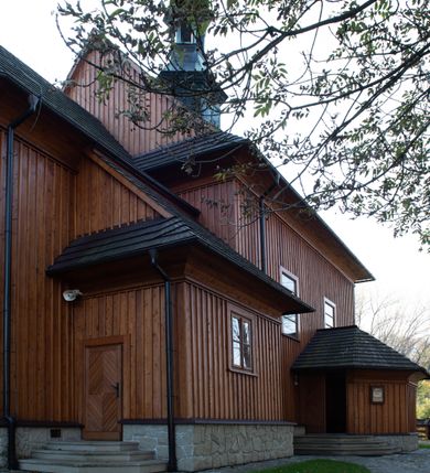 Zdjęcie nr 39: Kościół drewniany, orientowany, jednonawowy, wzniesiony na niewielkiej ceglanej podmurówce, o konstrukcji zrębowej, ściany szalowane pionowo z listowaniem, kruchta zachodnia o konstrukcji słupowo-ramowej. Nawa na rzucie prostokąta zbliżonego do kwadratu, prezbiterium węższe od nawy, wydłużone, zamknięte trójbocznie. Do północnej ściany prezbiterium przylega zakrystia na planie prostokąta. Do północnej ściany nawy przywiera niewielka kruchta na planie prostokąta, zamknięta trójbocznie. Przy zachodniej ścianie nawy znajduje się kruchta na rzucie prostokąta. Wnętrze nakryte pozornymi, spłaszczonymi sklepieniami o krzywiźnie przybierającej kształt łuku odcinkowego. Pomiędzy nawą a prezbiterium otwór tęczowy o wykroju prostokąta zbliżonego do kwadratu zamkniętego łukiem koszowym. Prezbiterium niższe od nawy, sięgające wysokości łuku tęczowego. Ściany prezbiterium i nawy obiega profilowany gzyms, oddzielający je od sklepień. W części zachodniej nawy parapet chóru muzycznego wsparty na dwóch kolumnach o stylizowanych kapitelach kompozytowych i kanelowanych trzonach na wysokich cokołach. W północno-zachodnim narożu drabiniaste schody prowadzące na chór w ośmiobocznej obudowie. Ściany i sklepienia pokryte polichromią. Na ścianach wzdłużnych oraz na osi ściany zachodniej otwory drzwiowe o prostokątnym wykroju zamknięte drzwiami spągowymi z desek oraz płycinowymi, na północnej ścianie prezbiterium prostokątny otwór drzwiowy do zakrystii. Posadzka z płyt kamiennych, szachownicowa. Kruchty i zakrystia nakryte stropami, przez nie dostęp do wnętrza. 
Elewacja frontowa jednoosiowa, zamknięta trójkątnym szczytem, poprzedzona kruchtą. Na osi wejście do kruchty zachodniej w postaci dwuskrzydłowych drzwi w formie stojącego prostokąta. Nad kruchtą pięterko dzwonowe z prostokątnym otworem zamykanym dwuskrzydłowymi okiennicami. Elewacja boczna południowa z czterema oknami, po dwa w nawie i prezbiterium. Elewacja północna przepruta dwoma oknami w części nawy. Okna krosnowe, w formie stojącego prostokąta, o niewielkich podziałach kwaterowych z witrażami. Na wschodniej ścianie kruchty północnej okno i jednoskrzydłowe drzwi. W zakrystii okno na ścianie północnej i drzwi jednoskrzydłowe na ścianie wschodniej. Kościół nakrywają dachy dwuspadowe, wyższy nad korpusem nawowym, niższy nad prezbiterium. Kruchta zachodnia w formie przysadzistej wieży, niższej od korpusu nawowego z pięterkiem dzwonowym, zwieńczonym hełmem baniastym, pokrytym gontem z iglicą oraz gałką i krzyżem. Kruchta północna z dachem wielospadowym. Zakrystia nakryta dachem pulpitowym. W połowie wysokości dachu znajduje się wieżyczka na sygnaturkę na ośmiobocznym obitym blachą trzonie, z ażurową latarnią nakrytą baniastym hełmem zakończonym kulą z krzyżem. 



