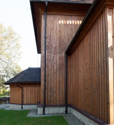 Zdjęcie nr 43: Kościół drewniany, orientowany, jednonawowy, wzniesiony na niewielkiej ceglanej podmurówce, o konstrukcji zrębowej, ściany szalowane pionowo z listowaniem, kruchta zachodnia o konstrukcji słupowo-ramowej. Nawa na rzucie prostokąta zbliżonego do kwadratu, prezbiterium węższe od nawy, wydłużone, zamknięte trójbocznie. Do północnej ściany prezbiterium przylega zakrystia na planie prostokąta. Do północnej ściany nawy przywiera niewielka kruchta na planie prostokąta, zamknięta trójbocznie. Przy zachodniej ścianie nawy znajduje się kruchta na rzucie prostokąta. Wnętrze nakryte pozornymi, spłaszczonymi sklepieniami o krzywiźnie przybierającej kształt łuku odcinkowego. Pomiędzy nawą a prezbiterium otwór tęczowy o wykroju prostokąta zbliżonego do kwadratu zamkniętego łukiem koszowym. Prezbiterium niższe od nawy, sięgające wysokości łuku tęczowego. Ściany prezbiterium i nawy obiega profilowany gzyms, oddzielający je od sklepień. W części zachodniej nawy parapet chóru muzycznego wsparty na dwóch kolumnach o stylizowanych kapitelach kompozytowych i kanelowanych trzonach na wysokich cokołach. W północno-zachodnim narożu drabiniaste schody prowadzące na chór w ośmiobocznej obudowie. Ściany i sklepienia pokryte polichromią. Na ścianach wzdłużnych oraz na osi ściany zachodniej otwory drzwiowe o prostokątnym wykroju zamknięte drzwiami spągowymi z desek oraz płycinowymi, na północnej ścianie prezbiterium prostokątny otwór drzwiowy do zakrystii. Posadzka z płyt kamiennych, szachownicowa. Kruchty i zakrystia nakryte stropami, przez nie dostęp do wnętrza. 
Elewacja frontowa jednoosiowa, zamknięta trójkątnym szczytem, poprzedzona kruchtą. Na osi wejście do kruchty zachodniej w postaci dwuskrzydłowych drzwi w formie stojącego prostokąta. Nad kruchtą pięterko dzwonowe z prostokątnym otworem zamykanym dwuskrzydłowymi okiennicami. Elewacja boczna południowa z czterema oknami, po dwa w nawie i prezbiterium. Elewacja północna przepruta dwoma oknami w części nawy. Okna krosnowe, w formie stojącego prostokąta, o niewielkich podziałach kwaterowych z witrażami. Na wschodniej ścianie kruchty północnej okno i jednoskrzydłowe drzwi. W zakrystii okno na ścianie północnej i drzwi jednoskrzydłowe na ścianie wschodniej. Kościół nakrywają dachy dwuspadowe, wyższy nad korpusem nawowym, niższy nad prezbiterium. Kruchta zachodnia w formie przysadzistej wieży, niższej od korpusu nawowego z pięterkiem dzwonowym, zwieńczonym hełmem baniastym, pokrytym gontem z iglicą oraz gałką i krzyżem. Kruchta północna z dachem wielospadowym. Zakrystia nakryta dachem pulpitowym. W połowie wysokości dachu znajduje się wieżyczka na sygnaturkę na ośmiobocznym obitym blachą trzonie, z ażurową latarnią nakrytą baniastym hełmem zakończonym kulą z krzyżem. 



