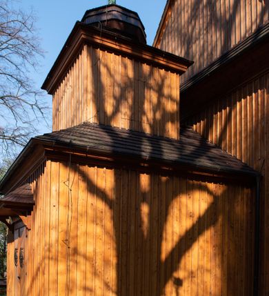 Zdjęcie nr 45: Kościół drewniany, orientowany, jednonawowy, wzniesiony na niewielkiej ceglanej podmurówce, o konstrukcji zrębowej, ściany szalowane pionowo z listowaniem, kruchta zachodnia o konstrukcji słupowo-ramowej. Nawa na rzucie prostokąta zbliżonego do kwadratu, prezbiterium węższe od nawy, wydłużone, zamknięte trójbocznie. Do północnej ściany prezbiterium przylega zakrystia na planie prostokąta. Do północnej ściany nawy przywiera niewielka kruchta na planie prostokąta, zamknięta trójbocznie. Przy zachodniej ścianie nawy znajduje się kruchta na rzucie prostokąta. Wnętrze nakryte pozornymi, spłaszczonymi sklepieniami o krzywiźnie przybierającej kształt łuku odcinkowego. Pomiędzy nawą a prezbiterium otwór tęczowy o wykroju prostokąta zbliżonego do kwadratu zamkniętego łukiem koszowym. Prezbiterium niższe od nawy, sięgające wysokości łuku tęczowego. Ściany prezbiterium i nawy obiega profilowany gzyms, oddzielający je od sklepień. W części zachodniej nawy parapet chóru muzycznego wsparty na dwóch kolumnach o stylizowanych kapitelach kompozytowych i kanelowanych trzonach na wysokich cokołach. W północno-zachodnim narożu drabiniaste schody prowadzące na chór w ośmiobocznej obudowie. Ściany i sklepienia pokryte polichromią. Na ścianach wzdłużnych oraz na osi ściany zachodniej otwory drzwiowe o prostokątnym wykroju zamknięte drzwiami spągowymi z desek oraz płycinowymi, na północnej ścianie prezbiterium prostokątny otwór drzwiowy do zakrystii. Posadzka z płyt kamiennych, szachownicowa. Kruchty i zakrystia nakryte stropami, przez nie dostęp do wnętrza. 
Elewacja frontowa jednoosiowa, zamknięta trójkątnym szczytem, poprzedzona kruchtą. Na osi wejście do kruchty zachodniej w postaci dwuskrzydłowych drzwi w formie stojącego prostokąta. Nad kruchtą pięterko dzwonowe z prostokątnym otworem zamykanym dwuskrzydłowymi okiennicami. Elewacja boczna południowa z czterema oknami, po dwa w nawie i prezbiterium. Elewacja północna przepruta dwoma oknami w części nawy. Okna krosnowe, w formie stojącego prostokąta, o niewielkich podziałach kwaterowych z witrażami. Na wschodniej ścianie kruchty północnej okno i jednoskrzydłowe drzwi. W zakrystii okno na ścianie północnej i drzwi jednoskrzydłowe na ścianie wschodniej. Kościół nakrywają dachy dwuspadowe, wyższy nad korpusem nawowym, niższy nad prezbiterium. Kruchta zachodnia w formie przysadzistej wieży, niższej od korpusu nawowego z pięterkiem dzwonowym, zwieńczonym hełmem baniastym, pokrytym gontem z iglicą oraz gałką i krzyżem. Kruchta północna z dachem wielospadowym. Zakrystia nakryta dachem pulpitowym. W połowie wysokości dachu znajduje się wieżyczka na sygnaturkę na ośmiobocznym obitym blachą trzonie, z ażurową latarnią nakrytą baniastym hełmem zakończonym kulą z krzyżem. 



