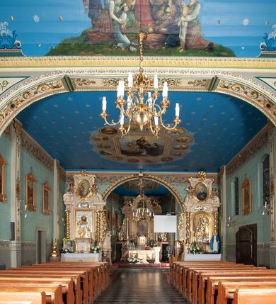 Zdjęcie nr 24: Kościół drewniany, orientowany, z dwuczęściowym korpusem: od wschodu jednonawowym, od zachodu trójnawowym; z prezbiterium zamkniętym trójbocznie, przy którym zakrystia od północy, z przybudówką od południa i dwiema wieżami od zachodu. Od wschodu chór muzyczny wbudowany pomiędzy wieże. Kościół wzniesiono w konstrukcji zrębowej, wieże w słupowo-ramowej, ściany oszalowane pionowym listwowaniem. 
Ściany prezbiterium i wschodniej części korpusu podzielone pseudokolumnami, zachodnia część podzielona kolumnami dźwigającymi architraw. Prezbiterium oddziela półokrągły łuk tęczowy; wschodnią i zachodnią część nawy łuk spłaszczony. Wschodnią część nawy oraz nawy boczne części zachodniej przekrywa płaski strop, zachodnią pozorne sklepienie zwierciadlane. Ściany oraz stropy pokryte polichromią figuralną, ornamentalną oraz roślinną: w prezbiterium namalowano wizerunek Ukrzyżowanego Chrystusa, przed łukiem tęczowym Matkę Boską w otoczeniu tajemnic różańcowych. Strop nawy imituje gwiaździste niebo. Posadzka kamienna. Wejście główne od zachodu, dwoje drzwi spod wież, kolejne od południa, przez przedsionek (babiniec), do zakrystii prowadzi portal zamknięty łukiem nadwieszonym. Cztery okna w prezbiterium o wykroju prostokątnym zamkniętym łukiem nadwieszonym, cztery we wschodniej części nawy o takim samym kształcie, cztery w zachodniej części zamknięte podwójnym łukiem kotarowym, dwa w zakrystii, południowej przybudówce, po dwa wysokie, wąskie okna w wieżach. Fasada dwuwieżowa, trójosiowa, trójkondygnacyjna. Przed wejściem kruchta zwieńczona dachem dwuspadowym, do której prowadzą dwuskrzydłowe drzwi w kształcie prostokątów, ściany boczne przeprute oknami. Powyżej dwie gładkie kondygnacje przedzielone dachem pulpitowym. Wieże trójkondygnacyjne, w dolnej kondygnacji drzwi, w wyższej okna w kształcie stojących prostokątów zamkniętych łukiem ostrym z wyciętym od dołu fragmentem pięcioliścia, powyżej ażurowa koronka. Ostatnią kondygnację tworzy izbica z nadwieszonym gankiem zamkniętym łukami spłaszczonymi. Od zewnątrz wyraźny podział na bryły, ściany gładkie. Każdą część przykrywa gontowy dach dwuspadowy, wspólny dla prezbiterium i starszej części, osobny dla nowszej części nawy; wieże nakryte hełmami namiotowymi, na kalenicy nawy baniasta wieżyczka na sygnaturkę z latarnią.  