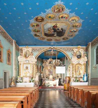 Zdjęcie nr 5: Kościół drewniany, orientowany, z dwuczęściowym korpusem: od wschodu jednonawowym, od zachodu trójnawowym; z prezbiterium zamkniętym trójbocznie, przy którym zakrystia od północy, z przybudówką od południa i dwiema wieżami od zachodu. Od wschodu chór muzyczny wbudowany pomiędzy wieże. Kościół wzniesiono w konstrukcji zrębowej, wieże w słupowo-ramowej, ściany oszalowane pionowym listwowaniem. 
Ściany prezbiterium i wschodniej części korpusu podzielone pseudokolumnami, zachodnia część podzielona kolumnami dźwigającymi architraw. Prezbiterium oddziela półokrągły łuk tęczowy; wschodnią i zachodnią część nawy łuk spłaszczony. Wschodnią część nawy oraz nawy boczne części zachodniej przekrywa płaski strop, zachodnią pozorne sklepienie zwierciadlane. Ściany oraz stropy pokryte polichromią figuralną, ornamentalną oraz roślinną: w prezbiterium namalowano wizerunek Ukrzyżowanego Chrystusa, przed łukiem tęczowym Matkę Boską w otoczeniu tajemnic różańcowych. Strop nawy imituje gwiaździste niebo. Posadzka kamienna. Wejście główne od zachodu, dwoje drzwi spod wież, kolejne od południa, przez przedsionek (babiniec), do zakrystii prowadzi portal zamknięty łukiem nadwieszonym. Cztery okna w prezbiterium o wykroju prostokątnym zamkniętym łukiem nadwieszonym, cztery we wschodniej części nawy o takim samym kształcie, cztery w zachodniej części zamknięte podwójnym łukiem kotarowym, dwa w zakrystii, południowej przybudówce, po dwa wysokie, wąskie okna w wieżach. Fasada dwuwieżowa, trójosiowa, trójkondygnacyjna. Przed wejściem kruchta zwieńczona dachem dwuspadowym, do której prowadzą dwuskrzydłowe drzwi w kształcie prostokątów, ściany boczne przeprute oknami. Powyżej dwie gładkie kondygnacje przedzielone dachem pulpitowym. Wieże trójkondygnacyjne, w dolnej kondygnacji drzwi, w wyższej okna w kształcie stojących prostokątów zamkniętych łukiem ostrym z wyciętym od dołu fragmentem pięcioliścia, powyżej ażurowa koronka. Ostatnią kondygnację tworzy izbica z nadwieszonym gankiem zamkniętym łukami spłaszczonymi. Od zewnątrz wyraźny podział na bryły, ściany gładkie. Każdą część przykrywa gontowy dach dwuspadowy, wspólny dla prezbiterium i starszej części, osobny dla nowszej części nawy; wieże nakryte hełmami namiotowymi, na kalenicy nawy baniasta wieżyczka na sygnaturkę z latarnią.  