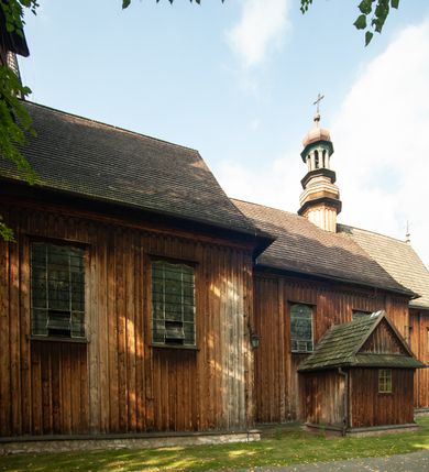Zdjęcie nr 20: Kościół drewniany, orientowany, z dwuczęściowym korpusem: od wschodu jednonawowym, od zachodu trójnawowym; z prezbiterium zamkniętym trójbocznie, przy którym zakrystia od północy, z przybudówką od południa i dwiema wieżami od zachodu. Od wschodu chór muzyczny wbudowany pomiędzy wieże. Kościół wzniesiono w konstrukcji zrębowej, wieże w słupowo-ramowej, ściany oszalowane pionowym listwowaniem. 
Ściany prezbiterium i wschodniej części korpusu podzielone pseudokolumnami, zachodnia część podzielona kolumnami dźwigającymi architraw. Prezbiterium oddziela półokrągły łuk tęczowy; wschodnią i zachodnią część nawy łuk spłaszczony. Wschodnią część nawy oraz nawy boczne części zachodniej przekrywa płaski strop, zachodnią pozorne sklepienie zwierciadlane. Ściany oraz stropy pokryte polichromią figuralną, ornamentalną oraz roślinną: w prezbiterium namalowano wizerunek Ukrzyżowanego Chrystusa, przed łukiem tęczowym Matkę Boską w otoczeniu tajemnic różańcowych. Strop nawy imituje gwiaździste niebo. Posadzka kamienna. Wejście główne od zachodu, dwoje drzwi spod wież, kolejne od południa, przez przedsionek (babiniec), do zakrystii prowadzi portal zamknięty łukiem nadwieszonym. Cztery okna w prezbiterium o wykroju prostokątnym zamkniętym łukiem nadwieszonym, cztery we wschodniej części nawy o takim samym kształcie, cztery w zachodniej części zamknięte podwójnym łukiem kotarowym, dwa w zakrystii, południowej przybudówce, po dwa wysokie, wąskie okna w wieżach. Fasada dwuwieżowa, trójosiowa, trójkondygnacyjna. Przed wejściem kruchta zwieńczona dachem dwuspadowym, do której prowadzą dwuskrzydłowe drzwi w kształcie prostokątów, ściany boczne przeprute oknami. Powyżej dwie gładkie kondygnacje przedzielone dachem pulpitowym. Wieże trójkondygnacyjne, w dolnej kondygnacji drzwi, w wyższej okna w kształcie stojących prostokątów zamkniętych łukiem ostrym z wyciętym od dołu fragmentem pięcioliścia, powyżej ażurowa koronka. Ostatnią kondygnację tworzy izbica z nadwieszonym gankiem zamkniętym łukami spłaszczonymi. Od zewnątrz wyraźny podział na bryły, ściany gładkie. Każdą część przykrywa gontowy dach dwuspadowy, wspólny dla prezbiterium i starszej części, osobny dla nowszej części nawy; wieże nakryte hełmami namiotowymi, na kalenicy nawy baniasta wieżyczka na sygnaturkę z latarnią.  