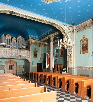 Zdjęcie nr 10: Kościół drewniany, orientowany, z dwuczęściowym korpusem: od wschodu jednonawowym, od zachodu trójnawowym; z prezbiterium zamkniętym trójbocznie, przy którym zakrystia od północy, z przybudówką od południa i dwiema wieżami od zachodu. Od wschodu chór muzyczny wbudowany pomiędzy wieże. Kościół wzniesiono w konstrukcji zrębowej, wieże w słupowo-ramowej, ściany oszalowane pionowym listwowaniem. 
Ściany prezbiterium i wschodniej części korpusu podzielone pseudokolumnami, zachodnia część podzielona kolumnami dźwigającymi architraw. Prezbiterium oddziela półokrągły łuk tęczowy; wschodnią i zachodnią część nawy łuk spłaszczony. Wschodnią część nawy oraz nawy boczne części zachodniej przekrywa płaski strop, zachodnią pozorne sklepienie zwierciadlane. Ściany oraz stropy pokryte polichromią figuralną, ornamentalną oraz roślinną: w prezbiterium namalowano wizerunek Ukrzyżowanego Chrystusa, przed łukiem tęczowym Matkę Boską w otoczeniu tajemnic różańcowych. Strop nawy imituje gwiaździste niebo. Posadzka kamienna. Wejście główne od zachodu, dwoje drzwi spod wież, kolejne od południa, przez przedsionek (babiniec), do zakrystii prowadzi portal zamknięty łukiem nadwieszonym. Cztery okna w prezbiterium o wykroju prostokątnym zamkniętym łukiem nadwieszonym, cztery we wschodniej części nawy o takim samym kształcie, cztery w zachodniej części zamknięte podwójnym łukiem kotarowym, dwa w zakrystii, południowej przybudówce, po dwa wysokie, wąskie okna w wieżach. Fasada dwuwieżowa, trójosiowa, trójkondygnacyjna. Przed wejściem kruchta zwieńczona dachem dwuspadowym, do której prowadzą dwuskrzydłowe drzwi w kształcie prostokątów, ściany boczne przeprute oknami. Powyżej dwie gładkie kondygnacje przedzielone dachem pulpitowym. Wieże trójkondygnacyjne, w dolnej kondygnacji drzwi, w wyższej okna w kształcie stojących prostokątów zamkniętych łukiem ostrym z wyciętym od dołu fragmentem pięcioliścia, powyżej ażurowa koronka. Ostatnią kondygnację tworzy izbica z nadwieszonym gankiem zamkniętym łukami spłaszczonymi. Od zewnątrz wyraźny podział na bryły, ściany gładkie. Każdą część przykrywa gontowy dach dwuspadowy, wspólny dla prezbiterium i starszej części, osobny dla nowszej części nawy; wieże nakryte hełmami namiotowymi, na kalenicy nawy baniasta wieżyczka na sygnaturkę z latarnią.  