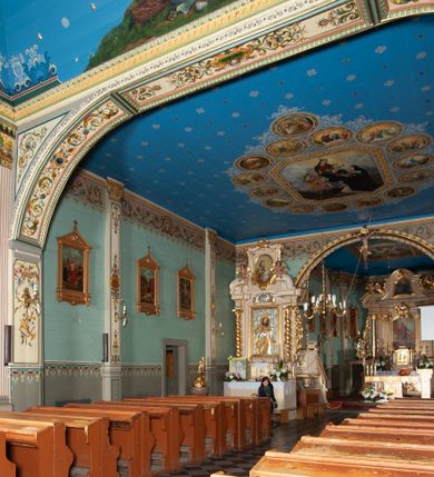 Zdjęcie nr 6: Kościół drewniany, orientowany, z dwuczęściowym korpusem: od wschodu jednonawowym, od zachodu trójnawowym; z prezbiterium zamkniętym trójbocznie, przy którym zakrystia od północy, z przybudówką od południa i dwiema wieżami od zachodu. Od wschodu chór muzyczny wbudowany pomiędzy wieże. Kościół wzniesiono w konstrukcji zrębowej, wieże w słupowo-ramowej, ściany oszalowane pionowym listwowaniem. 
Ściany prezbiterium i wschodniej części korpusu podzielone pseudokolumnami, zachodnia część podzielona kolumnami dźwigającymi architraw. Prezbiterium oddziela półokrągły łuk tęczowy; wschodnią i zachodnią część nawy łuk spłaszczony. Wschodnią część nawy oraz nawy boczne części zachodniej przekrywa płaski strop, zachodnią pozorne sklepienie zwierciadlane. Ściany oraz stropy pokryte polichromią figuralną, ornamentalną oraz roślinną: w prezbiterium namalowano wizerunek Ukrzyżowanego Chrystusa, przed łukiem tęczowym Matkę Boską w otoczeniu tajemnic różańcowych. Strop nawy imituje gwiaździste niebo. Posadzka kamienna. Wejście główne od zachodu, dwoje drzwi spod wież, kolejne od południa, przez przedsionek (babiniec), do zakrystii prowadzi portal zamknięty łukiem nadwieszonym. Cztery okna w prezbiterium o wykroju prostokątnym zamkniętym łukiem nadwieszonym, cztery we wschodniej części nawy o takim samym kształcie, cztery w zachodniej części zamknięte podwójnym łukiem kotarowym, dwa w zakrystii, południowej przybudówce, po dwa wysokie, wąskie okna w wieżach. Fasada dwuwieżowa, trójosiowa, trójkondygnacyjna. Przed wejściem kruchta zwieńczona dachem dwuspadowym, do której prowadzą dwuskrzydłowe drzwi w kształcie prostokątów, ściany boczne przeprute oknami. Powyżej dwie gładkie kondygnacje przedzielone dachem pulpitowym. Wieże trójkondygnacyjne, w dolnej kondygnacji drzwi, w wyższej okna w kształcie stojących prostokątów zamkniętych łukiem ostrym z wyciętym od dołu fragmentem pięcioliścia, powyżej ażurowa koronka. Ostatnią kondygnację tworzy izbica z nadwieszonym gankiem zamkniętym łukami spłaszczonymi. Od zewnątrz wyraźny podział na bryły, ściany gładkie. Każdą część przykrywa gontowy dach dwuspadowy, wspólny dla prezbiterium i starszej części, osobny dla nowszej części nawy; wieże nakryte hełmami namiotowymi, na kalenicy nawy baniasta wieżyczka na sygnaturkę z latarnią.  