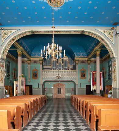 Zdjęcie nr 12: Kościół drewniany, orientowany, z dwuczęściowym korpusem: od wschodu jednonawowym, od zachodu trójnawowym; z prezbiterium zamkniętym trójbocznie, przy którym zakrystia od północy, z przybudówką od południa i dwiema wieżami od zachodu. Od wschodu chór muzyczny wbudowany pomiędzy wieże. Kościół wzniesiono w konstrukcji zrębowej, wieże w słupowo-ramowej, ściany oszalowane pionowym listwowaniem. 
Ściany prezbiterium i wschodniej części korpusu podzielone pseudokolumnami, zachodnia część podzielona kolumnami dźwigającymi architraw. Prezbiterium oddziela półokrągły łuk tęczowy; wschodnią i zachodnią część nawy łuk spłaszczony. Wschodnią część nawy oraz nawy boczne części zachodniej przekrywa płaski strop, zachodnią pozorne sklepienie zwierciadlane. Ściany oraz stropy pokryte polichromią figuralną, ornamentalną oraz roślinną: w prezbiterium namalowano wizerunek Ukrzyżowanego Chrystusa, przed łukiem tęczowym Matkę Boską w otoczeniu tajemnic różańcowych. Strop nawy imituje gwiaździste niebo. Posadzka kamienna. Wejście główne od zachodu, dwoje drzwi spod wież, kolejne od południa, przez przedsionek (babiniec), do zakrystii prowadzi portal zamknięty łukiem nadwieszonym. Cztery okna w prezbiterium o wykroju prostokątnym zamkniętym łukiem nadwieszonym, cztery we wschodniej części nawy o takim samym kształcie, cztery w zachodniej części zamknięte podwójnym łukiem kotarowym, dwa w zakrystii, południowej przybudówce, po dwa wysokie, wąskie okna w wieżach. Fasada dwuwieżowa, trójosiowa, trójkondygnacyjna. Przed wejściem kruchta zwieńczona dachem dwuspadowym, do której prowadzą dwuskrzydłowe drzwi w kształcie prostokątów, ściany boczne przeprute oknami. Powyżej dwie gładkie kondygnacje przedzielone dachem pulpitowym. Wieże trójkondygnacyjne, w dolnej kondygnacji drzwi, w wyższej okna w kształcie stojących prostokątów zamkniętych łukiem ostrym z wyciętym od dołu fragmentem pięcioliścia, powyżej ażurowa koronka. Ostatnią kondygnację tworzy izbica z nadwieszonym gankiem zamkniętym łukami spłaszczonymi. Od zewnątrz wyraźny podział na bryły, ściany gładkie. Każdą część przykrywa gontowy dach dwuspadowy, wspólny dla prezbiterium i starszej części, osobny dla nowszej części nawy; wieże nakryte hełmami namiotowymi, na kalenicy nawy baniasta wieżyczka na sygnaturkę z latarnią.  