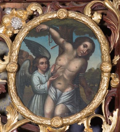 Zdjęcie nr 1: Obraz w kształcie owalu z przedstawieniem św. Sebastiana. W centrum kompozycji ukazany św. Sebastian w trzech czwartych wysokości, sylwetka esowato wygięta, głowa bezwładnie opuszczona na lewy bark, prawa ręka przymocowana do gałęzi wysoko nad głową, lewa ręka ugięta w łokciu, schowana za plecami świętego. Twarz o młodzieńczych rysach, zarumienionych policzkach, nos długi i wąski, głowa okolona bujnymi i ciemnymi włosami zasłaniającymi uszy. Święty z nagim torsem, przepasany w biodrach białą tkaniną. Z lewej strony stoi anioł rękami wskazujący na św. Sebastiana, ubrany w białą suknię z długimi rękawami i kołnierzykiem. W tle pejzaż i błękitne niebo, pośrodku wysokie drzewo. Kolorystyka ciepła.
