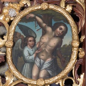 Zdjęcie nr 1: Obraz w kształcie owalu z przedstawieniem św. Sebastiana. W centrum kompozycji ukazany św. Sebastian w trzech czwartych wysokości, sylwetka esowato wygięta, głowa bezwładnie opuszczona na lewy bark, prawa ręka przymocowana do gałęzi wysoko nad głową, lewa ręka ugięta w łokciu, schowana za plecami świętego. Twarz o młodzieńczych rysach, zarumienionych policzkach, nos długi i wąski, głowa okolona bujnymi i ciemnymi włosami zasłaniającymi uszy. Święty z nagim torsem, przepasany w biodrach białą tkaniną. Z lewej strony stoi anioł rękami wskazujący na św. Sebastiana, ubrany w białą suknię z długimi rękawami i kołnierzykiem. W tle pejzaż i błękitne niebo, pośrodku wysokie drzewo. Kolorystyka ciepła.
