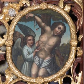 Zdjęcie nr 1: Obraz w kształcie owalu z przedstawieniem św. Sebastiana. W centrum kompozycji ukazany św. Sebastian w trzech czwartych wysokości, sylwetka esowato wygięta, głowa bezwładnie opuszczona na lewy bark, prawa ręka przymocowana do gałęzi wysoko nad głową, lewa ręka ugięta w łokciu, schowana za plecami świętego. Twarz o młodzieńczych rysach, zarumienionych policzkach, nos długi i wąski, głowa okolona bujnymi i ciemnymi włosami zasłaniającymi uszy. Święty z nagim torsem, przepasany w biodrach białą tkaniną. Z lewej strony stoi anioł rękami wskazujący na św. Sebastiana, ubrany w białą suknię z długimi rękawami i kołnierzykiem. W tle pejzaż i błękitne niebo, pośrodku wysokie drzewo. Kolorystyka ciepła.
