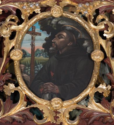 Zdjęcie nr 1: Obraz w kształcie owalu z przedstawieniem św. Franciszka z Asyżu. W centrum kompozycji święty ukazany w półpostaci, zwrócony delikatnie w prawo z dłońmi splecionymi i złożonymi na znajdującej się w dolnej części obrazu czaszce, głowa delikatnie uniesiona. Twarz podłużna, widoczna z lewego profilu, nos długi spiczasty, oczy ciemne i przymknięte, półkoliste łuki brwiowe, broda krótka i siwa. Na głowie tonsura, nad nią okrągły i złoty nimb. Święty jest ubrany w brązowy habit franciszkański. Na dłoniach oraz boku ma ślady po stygmatach. Po lewej stronie obrazu znajduje się krucyfiks. Krzyż brązowy i prosty. Chrystus rozpięty na wyprężonych ramionach, przybity do krzyża trzema gwoździami. Sylwetka szczupła, o ciemnej karnacji. Przewiązany jest w pasie białym perizonium, zawiązanym na prawym boku. Wokół głowy ma złoty i okrągły nimb. Nad nią wisi biały titulus w formie banderoli. W tle widoczny pejzaż z zabudowaniami oraz błękitno-różowe niebo, po prawej stronie obrazu ciemna szczelina skalna. Kolorystyka przygaszona. Rama złocona w formie wieńca z liści wawrzynu zdobiona czterema małymi różami.


