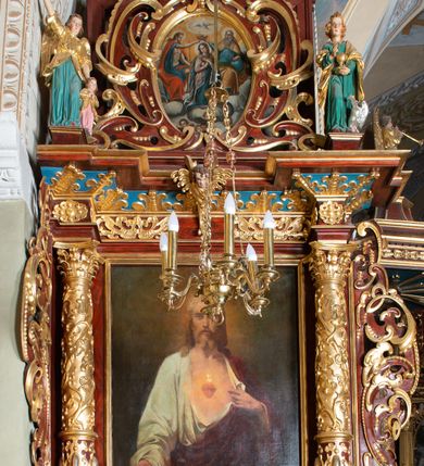 Zdjęcie nr 12: Ołtarz architektoniczny, jednoosiowy, jednokondygnacyjny ze zwieńczeniem. Ołtarz murowany, prostopadłościenny z kamienną mensą. Antepedium drewniane, w polu którego ozdobny, płaskorzeźbiony monogram maryjny na niebieskim tle oraz dekoracja ornamentalna w narożach pola. Pole ujęte ozdobną ramą z płaskorzeźbionym ornamentem z wici roślinnej na jasnoniebieskim tle. Drewniane retabulum ustawione na cokole. W predelli obraz z przedstawieniem Nawiedzenia Najświętszej Marii Panny w kształcie leżącego prostokąta o krótszych bokach zakończonych półkolistym łukiem nadwieszonym. Nastawa flankowana parą kolumn podtrzymujących wyłamane nad osią główną belkowanie. W polu głównym obraz Matki Boskiej Śnieżnej, a na zasuwie obraz Najświętsze Serce Jezusa. Zwieńczenie w formie owalu ujętego dekoracyjną, ażurową ramą formowaną z ornamentu małżowinowo-chrząstkowego na tle kwadratowej ramy. W polu znajduje się obraz „Koronacja Najświętszej Marii Panny”, a po bokach figury, po lewej: św. Archanioła Rafała z Tobiaszem, po prawej: św. Jana Ewangelisty, na szczycie figura św. Michała Archanioła. Partie cokołów ozdobione są ornamentem w formie kartusza o fantazyjnie kształtowanych krawędziach, z motywem kaboszonów pośrodku. Cokoły po zewnętrznej stronie zostały ozdobione ornamentalnym podwieszeniem z motywem małżowinowo-chrząstkowym. Zasadnicza część nastawy ujęta po bokach uszami z ornamentem o formach małżowinowo-chrząstkowych, z czego lewy złożony, dostosowany do północnego filaru kościoła. Trzony kolumn w dolnej partii ozdobione kartuszami o fantazyjnych wycięciach z motywami kaboszonów, w górnej partii pokryte dekoracją w formie wici winnej latorośli. W partii fryzu oraz pod gzymsem ornamentyka roślinna. Pośrodku belkowania motyw dekoracyjnego spływu z uskrzydloną główką aniołka z podwiązaną chustą pod szyją. Pole środkowe w formie prostokątnej wnęki ujętej profilowanymi ramami z czego wewnętrzna ozdobiona ornamentem małżowinowo-chrząstkowym z motywami uskrzydlonych główek aniołków pośrodku każdego boku ramy. Polichromia struktury ołtarza utrzymana w odcieniach ciemnego brązu i bordo, partie w tle ornamentów niebieskie. Ornamentyka i elementy detalu architektonicznego złocone i srebrzone. 