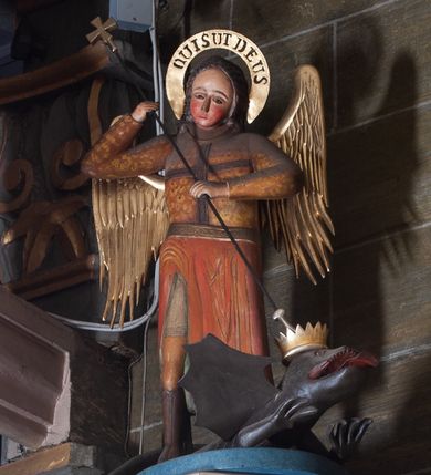 Zdjęcie nr 1: Ambona zawieszona na północnej ścianie prezbiterium. Kosz na rzucie sześcioboku (połowy ośmioboku), osadzony na nadwieszonej masywnej konsoli o formie odwróconego ściętego ostrosłupa o wklęsło-wypukłych bokach; kosz zwieńczony profilowanym gzymsem. Boki kosza ozdobione prostokątnymi płycinami w profilowanych obramieniach, w ich polach nałożone rzeźbione motywy rocaille’ów. Zaplecek w kształcie aediculi o wklęsło-wypukłych bokach, ujętej wolutami; w polu zaplecka płycina ozdobiona pozłoconym krzyżem łacińskim o trójdzielnych zakończeniach ramion i promieniami wokół przecięcia belek.  Baldachim na rzucie wieloboku opięty odcinkami występującego belkowania, ponad którymi woluty; wyżej ścięty ostrosłup o wklęsłych bokach tworzący podstawę dla pełnoplastycznej rzeźby św. Michała Archanioła przebijającego włócznią smoka leżącego u jego stóp. Na podniebiu podwieszona rzeźbiona Gołębica Ducha Świętego. Struktura pomalowana w kolorach ciemnego różu i ciemnego  niebieskiego, detale architektoniczne oraz motywy ornamentalne i rzeźbiarskie złocone lub srebrzone.