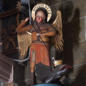 Zdjęcie nr 1: Ambona zawieszona na północnej ścianie prezbiterium. Kosz na rzucie sześcioboku (połowy ośmioboku), osadzony na nadwieszonej masywnej konsoli o formie odwróconego ściętego ostrosłupa o wklęsło-wypukłych bokach; kosz zwieńczony profilowanym gzymsem. Boki kosza ozdobione prostokątnymi płycinami w profilowanych obramieniach, w ich polach nałożone rzeźbione motywy rocaille’ów. Zaplecek w kształcie aediculi o wklęsło-wypukłych bokach, ujętej wolutami; w polu zaplecka płycina ozdobiona pozłoconym krzyżem łacińskim o trójdzielnych zakończeniach ramion i promieniami wokół przecięcia belek.  Baldachim na rzucie wieloboku opięty odcinkami występującego belkowania, ponad którymi woluty; wyżej ścięty ostrosłup o wklęsłych bokach tworzący podstawę dla pełnoplastycznej rzeźby św. Michała Archanioła przebijającego włócznią smoka leżącego u jego stóp. Na podniebiu podwieszona rzeźbiona Gołębica Ducha Świętego. Struktura pomalowana w kolorach ciemnego różu i ciemnego  niebieskiego, detale architektoniczne oraz motywy ornamentalne i rzeźbiarskie złocone lub srebrzone.