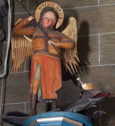 Zdjęcie nr 2: Ambona zawieszona na północnej ścianie prezbiterium. Kosz na rzucie sześcioboku (połowy ośmioboku), osadzony na nadwieszonej masywnej konsoli o formie odwróconego ściętego ostrosłupa o wklęsło-wypukłych bokach; kosz zwieńczony profilowanym gzymsem. Boki kosza ozdobione prostokątnymi płycinami w profilowanych obramieniach, w ich polach nałożone rzeźbione motywy rocaille’ów. Zaplecek w kształcie aediculi o wklęsło-wypukłych bokach, ujętej wolutami; w polu zaplecka płycina ozdobiona pozłoconym krzyżem łacińskim o trójdzielnych zakończeniach ramion i promieniami wokół przecięcia belek.  Baldachim na rzucie wieloboku opięty odcinkami występującego belkowania, ponad którymi woluty; wyżej ścięty ostrosłup o wklęsłych bokach tworzący podstawę dla pełnoplastycznej rzeźby św. Michała Archanioła przebijającego włócznią smoka leżącego u jego stóp. Na podniebiu podwieszona rzeźbiona Gołębica Ducha Świętego. Struktura pomalowana w kolorach ciemnego różu i ciemnego  niebieskiego, detale architektoniczne oraz motywy ornamentalne i rzeźbiarskie złocone lub srebrzone.