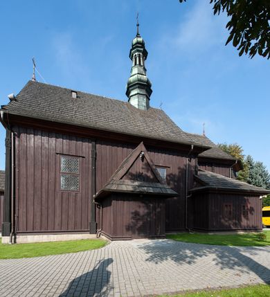 Zdjęcie nr 12: Kościół orientowany, jednonawowy, z węższym, zamkniętym wielobocznie prezbiterium, przy którym od północy prostokątna zakrystia (obecnie skarbiec) od południa prostokątna nowa zakrystia. Przy korpusie od południa i zachodu kruchty. Wewnątrz stropy, w prezbiterium i środkowej części nawy z fasetami; w nawie boczne płaskie odcinki stropu wparte na dwóch słupach. W prezbiterium i nawie na stropach dekoracja geometryczna z listew. Ściany artykułowane pilastrami. Tęcza o wystroju prostokątnym, ujęta przyściennymi filarami. Zewnątrz ściany szalowane deskowaniem na nakładkę, opięte pilastrami. Fasada rozczłonkowana czterema pilastrami wspierającymi gzyms, pośrodku wyłamany nad oknem wchodzącym w pole szczytu. Szczyt trójkątny, w połowie przedzielony gzymsem. Dachy dwuspadowe, nad prezbiterium wielopołaciowy, nad nową zakrystią namiotowy oraz pulpitowe; na kalenicy dachu nawy sygnaturka. Okna prostokątne w profilowanych obramieniach. Portal uszaty, z datą: 1748.