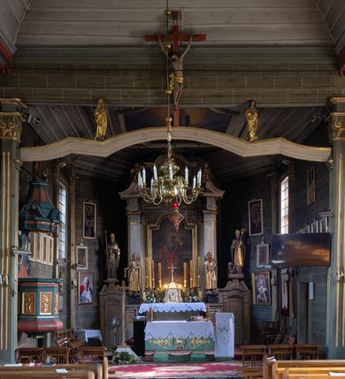 Zdjęcie nr 16: Kościół orientowany, jednonawowy, z węższym, zamkniętym wielobocznie prezbiterium, przy którym od północy prostokątna zakrystia (obecnie skarbiec) od południa prostokątna nowa zakrystia. Przy korpusie od południa i zachodu kruchty. Wewnątrz stropy, w prezbiterium i środkowej części nawy z fasetami; w nawie boczne płaskie odcinki stropu wparte na dwóch słupach. W prezbiterium i nawie na stropach dekoracja geometryczna z listew. Ściany artykułowane pilastrami. Tęcza o wystroju prostokątnym, ujęta przyściennymi filarami. Zewnątrz ściany szalowane deskowaniem na nakładkę, opięte pilastrami. Fasada rozczłonkowana czterema pilastrami wspierającymi gzyms, pośrodku wyłamany nad oknem wchodzącym w pole szczytu. Szczyt trójkątny, w połowie przedzielony gzymsem. Dachy dwuspadowe, nad prezbiterium wielopołaciowy, nad nową zakrystią namiotowy oraz pulpitowe; na kalenicy dachu nawy sygnaturka. Okna prostokątne w profilowanych obramieniach. Portal uszaty, z datą: 1748.