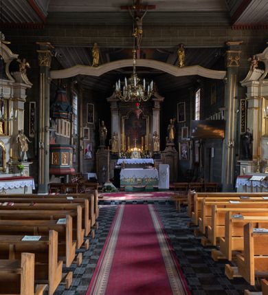 Zdjęcie nr 14: Kościół orientowany, jednonawowy, z węższym, zamkniętym wielobocznie prezbiterium, przy którym od północy prostokątna zakrystia (obecnie skarbiec) od południa prostokątna nowa zakrystia. Przy korpusie od południa i zachodu kruchty. Wewnątrz stropy, w prezbiterium i środkowej części nawy z fasetami; w nawie boczne płaskie odcinki stropu wparte na dwóch słupach. W prezbiterium i nawie na stropach dekoracja geometryczna z listew. Ściany artykułowane pilastrami. Tęcza o wystroju prostokątnym, ujęta przyściennymi filarami. Zewnątrz ściany szalowane deskowaniem na nakładkę, opięte pilastrami. Fasada rozczłonkowana czterema pilastrami wspierającymi gzyms, pośrodku wyłamany nad oknem wchodzącym w pole szczytu. Szczyt trójkątny, w połowie przedzielony gzymsem. Dachy dwuspadowe, nad prezbiterium wielopołaciowy, nad nową zakrystią namiotowy oraz pulpitowe; na kalenicy dachu nawy sygnaturka. Okna prostokątne w profilowanych obramieniach. Portal uszaty, z datą: 1748.