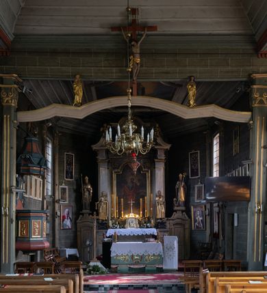 Zdjęcie nr 22: Kościół orientowany, jednonawowy, z węższym, zamkniętym wielobocznie prezbiterium, przy którym od północy prostokątna zakrystia (obecnie skarbiec) od południa prostokątna nowa zakrystia. Przy korpusie od południa i zachodu kruchty. Wewnątrz stropy, w prezbiterium i środkowej części nawy z fasetami; w nawie boczne płaskie odcinki stropu wparte na dwóch słupach. W prezbiterium i nawie na stropach dekoracja geometryczna z listew. Ściany artykułowane pilastrami. Tęcza o wystroju prostokątnym, ujęta przyściennymi filarami. Zewnątrz ściany szalowane deskowaniem na nakładkę, opięte pilastrami. Fasada rozczłonkowana czterema pilastrami wspierającymi gzyms, pośrodku wyłamany nad oknem wchodzącym w pole szczytu. Szczyt trójkątny, w połowie przedzielony gzymsem. Dachy dwuspadowe, nad prezbiterium wielopołaciowy, nad nową zakrystią namiotowy oraz pulpitowe; na kalenicy dachu nawy sygnaturka. Okna prostokątne w profilowanych obramieniach. Portal uszaty, z datą: 1748.
