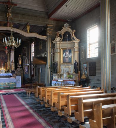 Zdjęcie nr 23: Kościół orientowany, jednonawowy, z węższym, zamkniętym wielobocznie prezbiterium, przy którym od północy prostokątna zakrystia (obecnie skarbiec) od południa prostokątna nowa zakrystia. Przy korpusie od południa i zachodu kruchty. Wewnątrz stropy, w prezbiterium i środkowej części nawy z fasetami; w nawie boczne płaskie odcinki stropu wparte na dwóch słupach. W prezbiterium i nawie na stropach dekoracja geometryczna z listew. Ściany artykułowane pilastrami. Tęcza o wystroju prostokątnym, ujęta przyściennymi filarami. Zewnątrz ściany szalowane deskowaniem na nakładkę, opięte pilastrami. Fasada rozczłonkowana czterema pilastrami wspierającymi gzyms, pośrodku wyłamany nad oknem wchodzącym w pole szczytu. Szczyt trójkątny, w połowie przedzielony gzymsem. Dachy dwuspadowe, nad prezbiterium wielopołaciowy, nad nową zakrystią namiotowy oraz pulpitowe; na kalenicy dachu nawy sygnaturka. Okna prostokątne w profilowanych obramieniach. Portal uszaty, z datą: 1748.