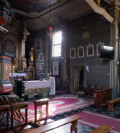 Zdjęcie nr 21: Kościół orientowany, jednonawowy, z węższym, zamkniętym wielobocznie prezbiterium, przy którym od północy prostokątna zakrystia (obecnie skarbiec) od południa prostokątna nowa zakrystia. Przy korpusie od południa i zachodu kruchty. Wewnątrz stropy, w prezbiterium i środkowej części nawy z fasetami; w nawie boczne płaskie odcinki stropu wparte na dwóch słupach. W prezbiterium i nawie na stropach dekoracja geometryczna z listew. Ściany artykułowane pilastrami. Tęcza o wystroju prostokątnym, ujęta przyściennymi filarami. Zewnątrz ściany szalowane deskowaniem na nakładkę, opięte pilastrami. Fasada rozczłonkowana czterema pilastrami wspierającymi gzyms, pośrodku wyłamany nad oknem wchodzącym w pole szczytu. Szczyt trójkątny, w połowie przedzielony gzymsem. Dachy dwuspadowe, nad prezbiterium wielopołaciowy, nad nową zakrystią namiotowy oraz pulpitowe; na kalenicy dachu nawy sygnaturka. Okna prostokątne w profilowanych obramieniach. Portal uszaty, z datą: 1748.