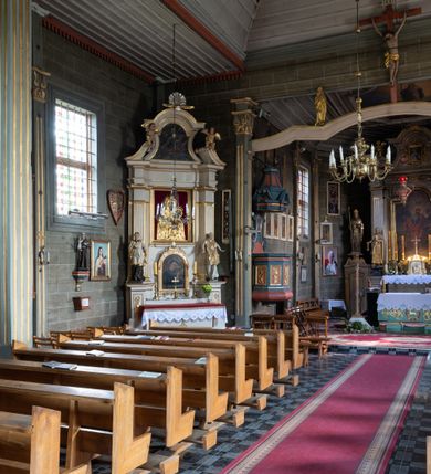 Zdjęcie nr 15: Kościół orientowany, jednonawowy, z węższym, zamkniętym wielobocznie prezbiterium, przy którym od północy prostokątna zakrystia (obecnie skarbiec) od południa prostokątna nowa zakrystia. Przy korpusie od południa i zachodu kruchty. Wewnątrz stropy, w prezbiterium i środkowej części nawy z fasetami; w nawie boczne płaskie odcinki stropu wparte na dwóch słupach. W prezbiterium i nawie na stropach dekoracja geometryczna z listew. Ściany artykułowane pilastrami. Tęcza o wystroju prostokątnym, ujęta przyściennymi filarami. Zewnątrz ściany szalowane deskowaniem na nakładkę, opięte pilastrami. Fasada rozczłonkowana czterema pilastrami wspierającymi gzyms, pośrodku wyłamany nad oknem wchodzącym w pole szczytu. Szczyt trójkątny, w połowie przedzielony gzymsem. Dachy dwuspadowe, nad prezbiterium wielopołaciowy, nad nową zakrystią namiotowy oraz pulpitowe; na kalenicy dachu nawy sygnaturka. Okna prostokątne w profilowanych obramieniach. Portal uszaty, z datą: 1748.