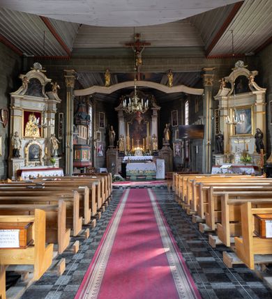 Zdjęcie nr 31: Kościół orientowany, jednonawowy, z węższym, zamkniętym wielobocznie prezbiterium, przy którym od północy prostokątna zakrystia (obecnie skarbiec) od południa prostokątna nowa zakrystia. Przy korpusie od południa i zachodu kruchty. Wewnątrz stropy, w prezbiterium i środkowej części nawy z fasetami; w nawie boczne płaskie odcinki stropu wparte na dwóch słupach. W prezbiterium i nawie na stropach dekoracja geometryczna z listew. Ściany artykułowane pilastrami. Tęcza o wystroju prostokątnym, ujęta przyściennymi filarami. Zewnątrz ściany szalowane deskowaniem na nakładkę, opięte pilastrami. Fasada rozczłonkowana czterema pilastrami wspierającymi gzyms, pośrodku wyłamany nad oknem wchodzącym w pole szczytu. Szczyt trójkątny, w połowie przedzielony gzymsem. Dachy dwuspadowe, nad prezbiterium wielopołaciowy, nad nową zakrystią namiotowy oraz pulpitowe; na kalenicy dachu nawy sygnaturka. Okna prostokątne w profilowanych obramieniach. Portal uszaty, z datą: 1748.