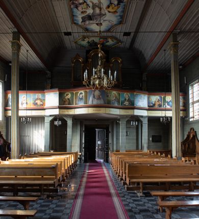 Zdjęcie nr 32: Kościół orientowany, jednonawowy, z węższym, zamkniętym wielobocznie prezbiterium, przy którym od północy prostokątna zakrystia (obecnie skarbiec) od południa prostokątna nowa zakrystia. Przy korpusie od południa i zachodu kruchty. Wewnątrz stropy, w prezbiterium i środkowej części nawy z fasetami; w nawie boczne płaskie odcinki stropu wparte na dwóch słupach. W prezbiterium i nawie na stropach dekoracja geometryczna z listew. Ściany artykułowane pilastrami. Tęcza o wystroju prostokątnym, ujęta przyściennymi filarami. Zewnątrz ściany szalowane deskowaniem na nakładkę, opięte pilastrami. Fasada rozczłonkowana czterema pilastrami wspierającymi gzyms, pośrodku wyłamany nad oknem wchodzącym w pole szczytu. Szczyt trójkątny, w połowie przedzielony gzymsem. Dachy dwuspadowe, nad prezbiterium wielopołaciowy, nad nową zakrystią namiotowy oraz pulpitowe; na kalenicy dachu nawy sygnaturka. Okna prostokątne w profilowanych obramieniach. Portal uszaty, z datą: 1748.