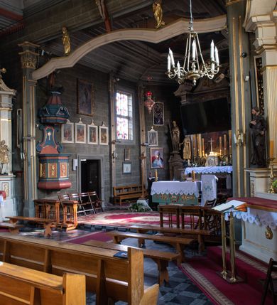 Zdjęcie nr 33: Kościół orientowany, jednonawowy, z węższym, zamkniętym wielobocznie prezbiterium, przy którym od północy prostokątna zakrystia (obecnie skarbiec) od południa prostokątna nowa zakrystia. Przy korpusie od południa i zachodu kruchty. Wewnątrz stropy, w prezbiterium i środkowej części nawy z fasetami; w nawie boczne płaskie odcinki stropu wparte na dwóch słupach. W prezbiterium i nawie na stropach dekoracja geometryczna z listew. Ściany artykułowane pilastrami. Tęcza o wystroju prostokątnym, ujęta przyściennymi filarami. Zewnątrz ściany szalowane deskowaniem na nakładkę, opięte pilastrami. Fasada rozczłonkowana czterema pilastrami wspierającymi gzyms, pośrodku wyłamany nad oknem wchodzącym w pole szczytu. Szczyt trójkątny, w połowie przedzielony gzymsem. Dachy dwuspadowe, nad prezbiterium wielopołaciowy, nad nową zakrystią namiotowy oraz pulpitowe; na kalenicy dachu nawy sygnaturka. Okna prostokątne w profilowanych obramieniach. Portal uszaty, z datą: 1748.