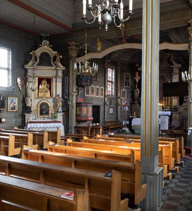 Zdjęcie nr 34: Kościół orientowany, jednonawowy, z węższym, zamkniętym wielobocznie prezbiterium, przy którym od północy prostokątna zakrystia (obecnie skarbiec) od południa prostokątna nowa zakrystia. Przy korpusie od południa i zachodu kruchty. Wewnątrz stropy, w prezbiterium i środkowej części nawy z fasetami; w nawie boczne płaskie odcinki stropu wparte na dwóch słupach. W prezbiterium i nawie na stropach dekoracja geometryczna z listew. Ściany artykułowane pilastrami. Tęcza o wystroju prostokątnym, ujęta przyściennymi filarami. Zewnątrz ściany szalowane deskowaniem na nakładkę, opięte pilastrami. Fasada rozczłonkowana czterema pilastrami wspierającymi gzyms, pośrodku wyłamany nad oknem wchodzącym w pole szczytu. Szczyt trójkątny, w połowie przedzielony gzymsem. Dachy dwuspadowe, nad prezbiterium wielopołaciowy, nad nową zakrystią namiotowy oraz pulpitowe; na kalenicy dachu nawy sygnaturka. Okna prostokątne w profilowanych obramieniach. Portal uszaty, z datą: 1748.
