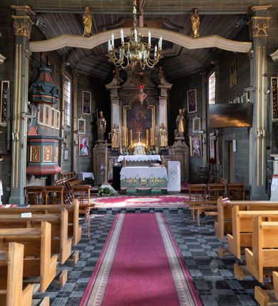 Zdjęcie nr 35: Kościół orientowany, jednonawowy, z węższym, zamkniętym wielobocznie prezbiterium, przy którym od północy prostokątna zakrystia (obecnie skarbiec) od południa prostokątna nowa zakrystia. Przy korpusie od południa i zachodu kruchty. Wewnątrz stropy, w prezbiterium i środkowej części nawy z fasetami; w nawie boczne płaskie odcinki stropu wparte na dwóch słupach. W prezbiterium i nawie na stropach dekoracja geometryczna z listew. Ściany artykułowane pilastrami. Tęcza o wystroju prostokątnym, ujęta przyściennymi filarami. Zewnątrz ściany szalowane deskowaniem na nakładkę, opięte pilastrami. Fasada rozczłonkowana czterema pilastrami wspierającymi gzyms, pośrodku wyłamany nad oknem wchodzącym w pole szczytu. Szczyt trójkątny, w połowie przedzielony gzymsem. Dachy dwuspadowe, nad prezbiterium wielopołaciowy, nad nową zakrystią namiotowy oraz pulpitowe; na kalenicy dachu nawy sygnaturka. Okna prostokątne w profilowanych obramieniach. Portal uszaty, z datą: 1748.