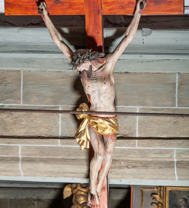 Zdjęcie nr 7: Rzeźby pełnoplastyczne, z tyłu opracowane, umieszczone na belce wygiętej łukiem wklęsło-wypukłym. Chrystus rozpięty na krzyżu w silnym zwisie ciała, z rękami skierowanymi niemal pionowo ku górze, z głową opuszczoną na prawy bark i nogami ugiętymi w kolanach, ze stopami założonymi lewa na prawą, przybitymi do krzyża jednym gwoździem. Biodra osłonięte perizonium ze zwisem tkaniny przy prawym udzie. Na belce krzyża ponad głową Chrystusa titulus z literami: I.N.R.I. Po prawej stronie Chrystusa Maria przedstawiona w całej postaci, zwrócona w trzech czwartych w lewo, w kierunku krzyża; stoi w lekkim kontrapoście, z prawą nogą ugiętą w kolanie, z rękami ugiętymi w łokciach i dłońmi na wysokości piersi. Ubrana w długą suknię z wąskimi rękawami, okryta wraz z głową długim do ziemi płaszczem układającym się sztywnymi, głęboko ciętymi fałdami. Po lewej stronie krzyża św. Jan Ewangelista przedstawiony w całej postaci, frontalnie do widza, stoi w lekkim kontrapoście, z lewą nogą ugiętą w kolanie, rękami ugiętymi w kolanie i dłońmi ułożonymi na wysokości piersi. Młodzieńcza twarz, bez zarostu, z lekko uniesionym podbródkiem i wzrokiem skierowanym ku górze, ujęta długimi, spływającymi na ramiona włosami. Ubrany w długą suknię z szerokimi, pofałdowanymi rękawami oraz zarzucony na ramiona płaszcz z prawą połą przerzuconą przez przód postaci na lewe przedramię i układający się na ziemi u stóp świętego. Karnacje jasne, Chrystusa z ranami, strużkami i kroplami krwi. 