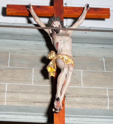 Zdjęcie nr 2: Rzeźby pełnoplastyczne, z tyłu opracowane, umieszczone na belce wygiętej łukiem wklęsło-wypukłym. Chrystus rozpięty na krzyżu w silnym zwisie ciała, z rękami skierowanymi niemal pionowo ku górze, z głową opuszczoną na prawy bark i nogami ugiętymi w kolanach, ze stopami założonymi lewa na prawą, przybitymi do krzyża jednym gwoździem. Biodra osłonięte perizonium ze zwisem tkaniny przy prawym udzie. Na belce krzyża ponad głową Chrystusa titulus z literami: I.N.R.I. Po prawej stronie Chrystusa Maria przedstawiona w całej postaci, zwrócona w trzech czwartych w lewo, w kierunku krzyża; stoi w lekkim kontrapoście, z prawą nogą ugiętą w kolanie, z rękami ugiętymi w łokciach i dłońmi na wysokości piersi. Ubrana w długą suknię z wąskimi rękawami, okryta wraz z głową długim do ziemi płaszczem układającym się sztywnymi, głęboko ciętymi fałdami. Po lewej stronie krzyża św. Jan Ewangelista przedstawiony w całej postaci, frontalnie do widza, stoi w lekkim kontrapoście, z lewą nogą ugiętą w kolanie, rękami ugiętymi w kolanie i dłońmi ułożonymi na wysokości piersi. Młodzieńcza twarz, bez zarostu, z lekko uniesionym podbródkiem i wzrokiem skierowanym ku górze, ujęta długimi, spływającymi na ramiona włosami. Ubrany w długą suknię z szerokimi, pofałdowanymi rękawami oraz zarzucony na ramiona płaszcz z prawą połą przerzuconą przez przód postaci na lewe przedramię i układający się na ziemi u stóp świętego. Karnacje jasne, Chrystusa z ranami, strużkami i kroplami krwi. 