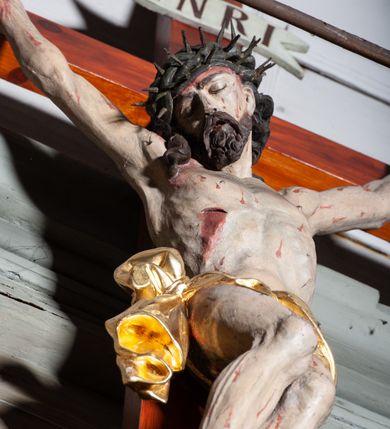 Zdjęcie nr 3: Rzeźby pełnoplastyczne, z tyłu opracowane, umieszczone na belce wygiętej łukiem wklęsło-wypukłym. Chrystus rozpięty na krzyżu w silnym zwisie ciała, z rękami skierowanymi niemal pionowo ku górze, z głową opuszczoną na prawy bark i nogami ugiętymi w kolanach, ze stopami założonymi lewa na prawą, przybitymi do krzyża jednym gwoździem. Biodra osłonięte perizonium ze zwisem tkaniny przy prawym udzie. Na belce krzyża ponad głową Chrystusa titulus z literami: I.N.R.I. Po prawej stronie Chrystusa Maria przedstawiona w całej postaci, zwrócona w trzech czwartych w lewo, w kierunku krzyża; stoi w lekkim kontrapoście, z prawą nogą ugiętą w kolanie, z rękami ugiętymi w łokciach i dłońmi na wysokości piersi. Ubrana w długą suknię z wąskimi rękawami, okryta wraz z głową długim do ziemi płaszczem układającym się sztywnymi, głęboko ciętymi fałdami. Po lewej stronie krzyża św. Jan Ewangelista przedstawiony w całej postaci, frontalnie do widza, stoi w lekkim kontrapoście, z lewą nogą ugiętą w kolanie, rękami ugiętymi w kolanie i dłońmi ułożonymi na wysokości piersi. Młodzieńcza twarz, bez zarostu, z lekko uniesionym podbródkiem i wzrokiem skierowanym ku górze, ujęta długimi, spływającymi na ramiona włosami. Ubrany w długą suknię z szerokimi, pofałdowanymi rękawami oraz zarzucony na ramiona płaszcz z prawą połą przerzuconą przez przód postaci na lewe przedramię i układający się na ziemi u stóp świętego. Karnacje jasne, Chrystusa z ranami, strużkami i kroplami krwi. 