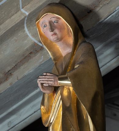 Zdjęcie nr 6: Rzeźby pełnoplastyczne, z tyłu opracowane, umieszczone na belce wygiętej łukiem wklęsło-wypukłym. Chrystus rozpięty na krzyżu w silnym zwisie ciała, z rękami skierowanymi niemal pionowo ku górze, z głową opuszczoną na prawy bark i nogami ugiętymi w kolanach, ze stopami założonymi lewa na prawą, przybitymi do krzyża jednym gwoździem. Biodra osłonięte perizonium ze zwisem tkaniny przy prawym udzie. Na belce krzyża ponad głową Chrystusa titulus z literami: I.N.R.I. Po prawej stronie Chrystusa Maria przedstawiona w całej postaci, zwrócona w trzech czwartych w lewo, w kierunku krzyża; stoi w lekkim kontrapoście, z prawą nogą ugiętą w kolanie, z rękami ugiętymi w łokciach i dłońmi na wysokości piersi. Ubrana w długą suknię z wąskimi rękawami, okryta wraz z głową długim do ziemi płaszczem układającym się sztywnymi, głęboko ciętymi fałdami. Po lewej stronie krzyża św. Jan Ewangelista przedstawiony w całej postaci, frontalnie do widza, stoi w lekkim kontrapoście, z lewą nogą ugiętą w kolanie, rękami ugiętymi w kolanie i dłońmi ułożonymi na wysokości piersi. Młodzieńcza twarz, bez zarostu, z lekko uniesionym podbródkiem i wzrokiem skierowanym ku górze, ujęta długimi, spływającymi na ramiona włosami. Ubrany w długą suknię z szerokimi, pofałdowanymi rękawami oraz zarzucony na ramiona płaszcz z prawą połą przerzuconą przez przód postaci na lewe przedramię i układający się na ziemi u stóp świętego. Karnacje jasne, Chrystusa z ranami, strużkami i kroplami krwi. 