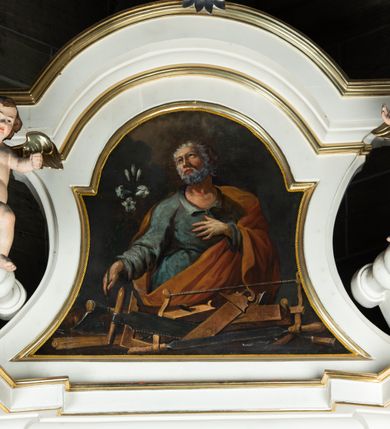 Zdjęcie nr 9: Obraz w kształcie prostokąta o wklęsłych bokach, zamkniętego łukiem półkolistym nadwieszonym, powtarzającym kształt zwieńczenia, z przedstawieniem św. Józefa w półpostaci. Święty ukazany jako starszy mężczyzna, frontalnie, z głową zwróconą lekko w prawo i wzrokiem skierowanym ku górze. Twarz pełna, z wysokim czołem i prostym nosem, okolona krótkimi kędzierzawymi, siwymi włosami i takimż zarostem. W prawej ręce, odwiedzionej od ciała nieco w bok i wyciągniętej do przodu, trzyma białą lilię, lewa ręka ugięta w łokciu, z dłonią przyłożoną do piersi. Ubrany w niebieską suknię z rozcięciem na piersi i szerokimi rękawami; okryty czerwonym płaszczem zarzuconym na lewe ramię, z prawą połą przełożoną przez przód postaci i przerzuconą przez lewe przedramię. Przed nim na pierwszym planie rozłożone narzędzia ciesielskie. W tle niebo i skłębione obłoki.  