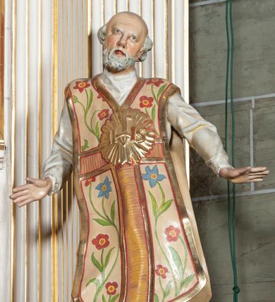 Zdjęcie nr 1: Rzeźba pełnoplastyczna, przyścienna; całopostaciowe przedstawienie św. Ignacego Loyoli, ukazanego frontalnie, stojącego w dynamicznej pozie, z esowato wygiętym ciałem, z prawą stopą wysuniętą do przodu, z rękami odwiedzionymi od ciała na boki, z głową przechyloną na lewy bark. Głowa starszego mężczyzny, z łysą czaszką, okolona wianuszkiem siwych włosów; twarz pociągła o jasnej karnacji, z zaróżowionymi policzkami, długim nosem i oczami skierowanymi ku górze, okolona obfitym siwym zarostem. Święty ubrany w długą albę ozdobioną u dołu koronką oraz ornat ozdobiony złotym galonem, bokami z motywem wici z barwnych kwiatów na białym tle i pretekstą w kształcie litery tau. Na piersiach okazały płaskorzeźbiony, kolisty złoty medalion z literami IHS w glorii promienistej. 