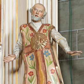 Zdjęcie nr 1: Rzeźba pełnoplastyczna, przyścienna; całopostaciowe przedstawienie św. Ignacego Loyoli, ukazanego frontalnie, stojącego w dynamicznej pozie, z esowato wygiętym ciałem, z prawą stopą wysuniętą do przodu, z rękami odwiedzionymi od ciała na boki, z głową przechyloną na lewy bark. Głowa starszego mężczyzny, z łysą czaszką, okolona wianuszkiem siwych włosów; twarz pociągła o jasnej karnacji, z zaróżowionymi policzkami, długim nosem i oczami skierowanymi ku górze, okolona obfitym siwym zarostem. Święty ubrany w długą albę ozdobioną u dołu koronką oraz ornat ozdobiony złotym galonem, bokami z motywem wici z barwnych kwiatów na białym tle i pretekstą w kształcie litery tau. Na piersiach okazały płaskorzeźbiony, kolisty złoty medalion z literami IHS w glorii promienistej. 