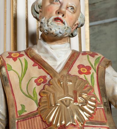 Zdjęcie nr 9: Rzeźba pełnoplastyczna, przyścienna; całopostaciowe przedstawienie św. Ignacego Loyoli, ukazanego frontalnie, stojącego w dynamicznej pozie, z esowato wygiętym ciałem, z prawą stopą wysuniętą do przodu, z rękami odwiedzionymi od ciała na boki, z głową przechyloną na lewy bark. Głowa starszego mężczyzny, z łysą czaszką, okolona wianuszkiem siwych włosów; twarz pociągła o jasnej karnacji, z zaróżowionymi policzkami, długim nosem i oczami skierowanymi ku górze, okolona obfitym siwym zarostem. Święty ubrany w długą albę ozdobioną u dołu koronką oraz ornat ozdobiony złotym galonem, bokami z motywem wici z barwnych kwiatów na białym tle i pretekstą w kształcie litery tau. Na piersiach okazały płaskorzeźbiony, kolisty złoty medalion z literami IHS w glorii promienistej. 