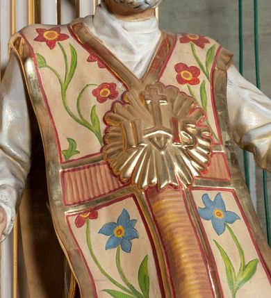 Zdjęcie nr 2: Rzeźba pełnoplastyczna, przyścienna; całopostaciowe przedstawienie św. Ignacego Loyoli, ukazanego frontalnie, stojącego w dynamicznej pozie, z esowato wygiętym ciałem, z prawą stopą wysuniętą do przodu, z rękami odwiedzionymi od ciała na boki, z głową przechyloną na lewy bark. Głowa starszego mężczyzny, z łysą czaszką, okolona wianuszkiem siwych włosów; twarz pociągła o jasnej karnacji, z zaróżowionymi policzkami, długim nosem i oczami skierowanymi ku górze, okolona obfitym siwym zarostem. Święty ubrany w długą albę ozdobioną u dołu koronką oraz ornat ozdobiony złotym galonem, bokami z motywem wici z barwnych kwiatów na białym tle i pretekstą w kształcie litery tau. Na piersiach okazały płaskorzeźbiony, kolisty złoty medalion z literami IHS w glorii promienistej. 