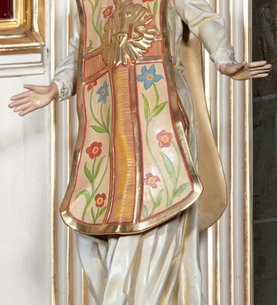 Zdjęcie nr 4: Rzeźba pełnoplastyczna, przyścienna; całopostaciowe przedstawienie św. Ignacego Loyoli, ukazanego frontalnie, stojącego w dynamicznej pozie, z esowato wygiętym ciałem, z prawą stopą wysuniętą do przodu, z rękami odwiedzionymi od ciała na boki, z głową przechyloną na lewy bark. Głowa starszego mężczyzny, z łysą czaszką, okolona wianuszkiem siwych włosów; twarz pociągła o jasnej karnacji, z zaróżowionymi policzkami, długim nosem i oczami skierowanymi ku górze, okolona obfitym siwym zarostem. Święty ubrany w długą albę ozdobioną u dołu koronką oraz ornat ozdobiony złotym galonem, bokami z motywem wici z barwnych kwiatów na białym tle i pretekstą w kształcie litery tau. Na piersiach okazały płaskorzeźbiony, kolisty złoty medalion z literami IHS w glorii promienistej. 