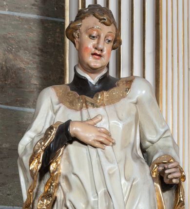 Zdjęcie nr 1: Rzeźba pełnoplastyczna, przyścienna; całopostaciowe przedstawienie św. Stanisława Kostki, ukazanego frontalnie, stojącego w ożywionej pozie, z esowato wygiętym ciałem, z prawą stopą wysuniętą do przodu, z lewą ręką wysuniętą do przodu i prawą dłonią złożoną na piersi, z głową lekko przechyloną na lewy bark., w; Twarz młodego mężczyzny o niemal jeszcze dziecięcych rysach, o jasnej karnacji, z zaróżowionymi policzkami, długim nosem i oczami skierowanymi w dół. Święty ubrany w udrapowane dynamicznie czarną sutannę jezuicką i białą komżę ozdobioną złotą koronką.