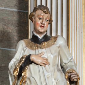 Zdjęcie nr 1: Rzeźba pełnoplastyczna, przyścienna; całopostaciowe przedstawienie św. Stanisława Kostki, ukazanego frontalnie, stojącego w ożywionej pozie, z esowato wygiętym ciałem, z prawą stopą wysuniętą do przodu, z lewą ręką wysuniętą do przodu i prawą dłonią złożoną na piersi, z głową lekko przechyloną na lewy bark., w; Twarz młodego mężczyzny o niemal jeszcze dziecięcych rysach, o jasnej karnacji, z zaróżowionymi policzkami, długim nosem i oczami skierowanymi w dół. Święty ubrany w udrapowane dynamicznie czarną sutannę jezuicką i białą komżę ozdobioną złotą koronką.