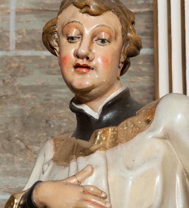 Zdjęcie nr 4: Rzeźba pełnoplastyczna, przyścienna; całopostaciowe przedstawienie św. Stanisława Kostki, ukazanego frontalnie, stojącego w ożywionej pozie, z esowato wygiętym ciałem, z prawą stopą wysuniętą do przodu, z lewą ręką wysuniętą do przodu i prawą dłonią złożoną na piersi, z głową lekko przechyloną na lewy bark., w; Twarz młodego mężczyzny o niemal jeszcze dziecięcych rysach, o jasnej karnacji, z zaróżowionymi policzkami, długim nosem i oczami skierowanymi w dół. Święty ubrany w udrapowane dynamicznie czarną sutannę jezuicką i białą komżę ozdobioną złotą koronką.