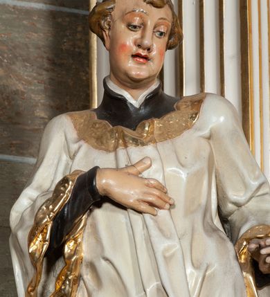 Zdjęcie nr 3: Rzeźba pełnoplastyczna, przyścienna; całopostaciowe przedstawienie św. Stanisława Kostki, ukazanego frontalnie, stojącego w ożywionej pozie, z esowato wygiętym ciałem, z prawą stopą wysuniętą do przodu, z lewą ręką wysuniętą do przodu i prawą dłonią złożoną na piersi, z głową lekko przechyloną na lewy bark., w; Twarz młodego mężczyzny o niemal jeszcze dziecięcych rysach, o jasnej karnacji, z zaróżowionymi policzkami, długim nosem i oczami skierowanymi w dół. Święty ubrany w udrapowane dynamicznie czarną sutannę jezuicką i białą komżę ozdobioną złotą koronką.