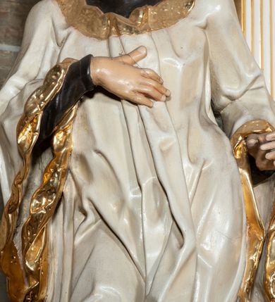 Zdjęcie nr 2: Rzeźba pełnoplastyczna, przyścienna; całopostaciowe przedstawienie św. Stanisława Kostki, ukazanego frontalnie, stojącego w ożywionej pozie, z esowato wygiętym ciałem, z prawą stopą wysuniętą do przodu, z lewą ręką wysuniętą do przodu i prawą dłonią złożoną na piersi, z głową lekko przechyloną na lewy bark., w; Twarz młodego mężczyzny o niemal jeszcze dziecięcych rysach, o jasnej karnacji, z zaróżowionymi policzkami, długim nosem i oczami skierowanymi w dół. Święty ubrany w udrapowane dynamicznie czarną sutannę jezuicką i białą komżę ozdobioną złotą koronką.