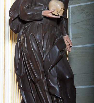 Zdjęcie nr 2: Rzeźba pełnoplastyczna, przyścienna ukazująca jezuickiego świętego św. Franciszka Borgiasza w całej postaci, w ujęciu frontalnym. Święty o szczupłej sylwetce stoi w kontrapoście, z lewą nogą ugiętą w kolanie i wysuniętą stopą, z głową przechyloną na prawy bark. Lewa ręka opuszczona wzdłuż tułowia, w prawej ugiętej w łokciu trzyma na wysokości piersi czaszkę ludzką. Twarz szczupła, o jasnej karnacji, z długim nosem i wzrokiem skierowanym d dół; włosy krótkie, zmierzwione. Ubrany w czarną sutannę jezuicką i płaszcz. 