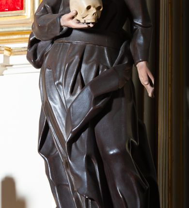 Zdjęcie nr 6: Rzeźba pełnoplastyczna, przyścienna ukazująca jezuickiego świętego św. Franciszka Borgiasza w całej postaci, w ujęciu frontalnym. Święty o szczupłej sylwetce stoi w kontrapoście, z lewą nogą ugiętą w kolanie i wysuniętą stopą, z głową przechyloną na prawy bark. Lewa ręka opuszczona wzdłuż tułowia, w prawej ugiętej w łokciu trzyma na wysokości piersi czaszkę ludzką. Twarz szczupła, o jasnej karnacji, z długim nosem i wzrokiem skierowanym d dół; włosy krótkie, zmierzwione. Ubrany w czarną sutannę jezuicką i płaszcz. 