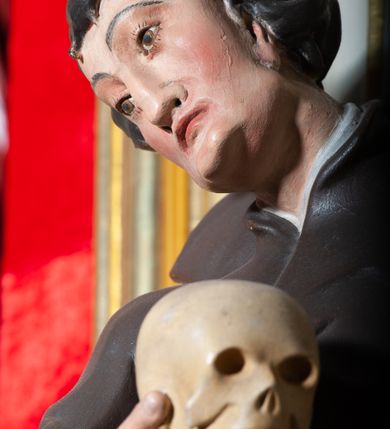 Zdjęcie nr 7: Rzeźba pełnoplastyczna, przyścienna ukazująca jezuickiego świętego św. Franciszka Borgiasza w całej postaci, w ujęciu frontalnym. Święty o szczupłej sylwetce stoi w kontrapoście, z lewą nogą ugiętą w kolanie i wysuniętą stopą, z głową przechyloną na prawy bark. Lewa ręka opuszczona wzdłuż tułowia, w prawej ugiętej w łokciu trzyma na wysokości piersi czaszkę ludzką. Twarz szczupła, o jasnej karnacji, z długim nosem i wzrokiem skierowanym d dół; włosy krótkie, zmierzwione. Ubrany w czarną sutannę jezuicką i płaszcz. 