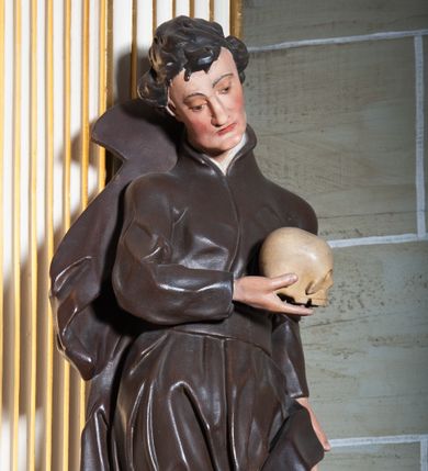 Zdjęcie nr 1: Rzeźba pełnoplastyczna, przyścienna ukazująca jezuickiego świętego św. Franciszka Borgiasza w całej postaci, w ujęciu frontalnym. Święty o szczupłej sylwetce stoi w kontrapoście, z lewą nogą ugiętą w kolanie i wysuniętą stopą, z głową przechyloną na prawy bark. Lewa ręka opuszczona wzdłuż tułowia, w prawej ugiętej w łokciu trzyma na wysokości piersi czaszkę ludzką. Twarz szczupła, o jasnej karnacji, z długim nosem i wzrokiem skierowanym d dół; włosy krótkie, zmierzwione. Ubrany w czarną sutannę jezuicką i płaszcz. 
