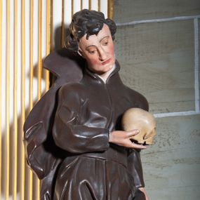 Zdjęcie nr 1: Rzeźba pełnoplastyczna, przyścienna ukazująca jezuickiego świętego św. Franciszka Borgiasza w całej postaci, w ujęciu frontalnym. Święty o szczupłej sylwetce stoi w kontrapoście, z lewą nogą ugiętą w kolanie i wysuniętą stopą, z głową przechyloną na prawy bark. Lewa ręka opuszczona wzdłuż tułowia, w prawej ugiętej w łokciu trzyma na wysokości piersi czaszkę ludzką. Twarz szczupła, o jasnej karnacji, z długim nosem i wzrokiem skierowanym d dół; włosy krótkie, zmierzwione. Ubrany w czarną sutannę jezuicką i płaszcz. 